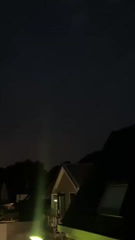 The video shows a nighttime view of rooftops and trees under a dark sky. In the second frame, a bright flash of lightning illuminates the sky and a portion of the trees. No people are visible.