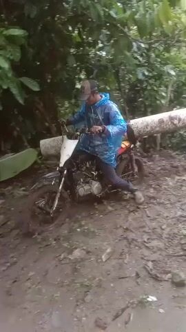 A man wearing a blue plastic poncho and a baseball cap drives a motorcycle along a muddy path. A log is strapped horizontally across the back of the bike. The first part of the video is filmed in the woods, then the path leads to a grassy area with houses. The man rides the motorcycle past a house with white pillars and a tiled porch.