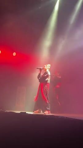 Two women are on a stage. One woman is in the foreground, holding a microphone and singing. She is wearing a black jacket with red fringe, black pants, and sneakers. The second woman is behind her and slightly to the right, also holding a microphone. She is wearing a black outfit with red straps. The stage is lit with pink and red lights, and there is smoke in the air. There is a banner on the left side of the stage with writing on it. The video is a close-up shot of the performers.