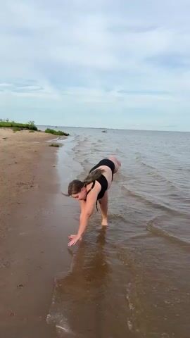 A woman in a black bikini is performing a handstand on a sandy beach. She is near the water's edge. The camera pans to the right, showing the woman in a crouched position in the shallow water. A dog walks into the frame from the right and stands near the woman's legs.