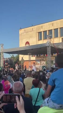 This is a video of an outdoor event taking place in Etropole, Bulgaria. A large crowd of people are gathered in front of a stage, where a performance is underway. On the stage, there are several performers, including a person in a white dress who appears to be singing, and a statue of a man standing behind them. A band with musical instruments is also visible on the stage. The event is being held in front of a large, pale building with a prominent red and white circular emblem on its facade. The sky is clear and blue, suggesting it is daytime, likely late afternoon or early evening given the warm lighting. Some attendees are holding up their mobile phones to record the performance, indicating engagement and interest. In the foreground, a pink inflatable balloon shaped like a cartoon character is visible, adding a festive touch to the scene. The overall atmosphere appears to be lively and celebratory, with a significant turnout of people enjoying the event.