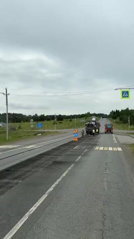 This video shows roadwork in progress on a cloudy day. A large asphalt milling machine is positioned on the right side of the road, with a dump truck connected to its rear. Several construction workers are present, some wearing high-visibility vests. Traffic cones and road signs, including a "men at work" sign and a directional arrow sign, are placed on the left side of the road. The road surface in front of the milling machine appears to have been recently worked on, with loose asphalt visible. A pedestrian crossing with yellow and white stripes is also visible on the road. The background shows a landscape with trees and fields, and power lines are overhead.