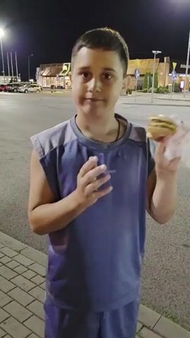 A young boy stands outside at night holding a hamburger and a white napkin. He takes a bite of the hamburger and chews it. In the background, a McDonald's restaurant is visible with its illuminated sign. The boy is wearing a dark blue tank top with grey trim. The ground is paved with bricks, and a street is visible behind him. Streetlights illuminate the scene.