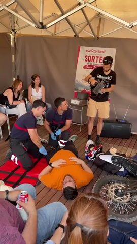 Here is a description of the scene in the video.

The scene is set outdoors, beneath a temporary shelter of some kind. Several people sit in chairs watching a demonstration. On the floor, a man in an orange shirt lies on his back. Two men in blue and red uniforms kneel at his side, appearing to attend to him. Beside them is a white motorcycle with a helmet lying near its back wheel.

A man in a black t-shirt and khaki shorts stands near the downed motorcycle, holding a microphone and appearing to address the gathered audience. A woman stands to his right, also holding a microphone. The man on the left side of the screen seems to be recording the demonstration. A boombox sits on a small table behind him.

A poster with the word “switzerland” is visible in the background.