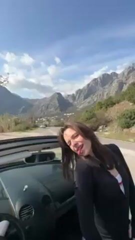 The video begins with a woman standing next to a convertible car. She is wearing a black jacket, and behind her is a mountain range, a winding road, and a town. The camera then shifts to focus on the mountains, which are rocky and sparsely covered with trees. The sky is blue with some clouds.
