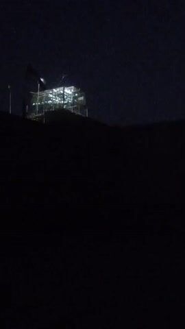 The video shows a dark, nighttime scene with a lit structure on a hill. The structure appears to be under construction or is a temporary setup, with scaffolding visible. Lights are illuminating the interior and exterior of the structure, creating a bright contrast against the dark sky and landscape. There is a flag visible at the top of the structure, flying from a pole. The foreground is mostly dark and indistinct, suggesting the viewpoint is from a distance or low on the hill. The overall impression is one of a remote or elevated location, possibly a construction site or a temporary installation, observed at night.
