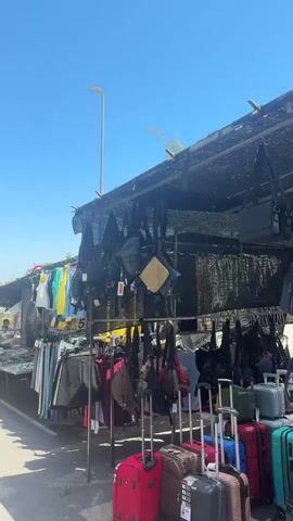 This media file captures a bustling outdoor market scene under a clear, bright blue sky, suggesting a sunny day, likely mid-morning or early afternoon due to the strong shadows. The setting appears to be a street lined with temporary market stalls, some covered by green and black canopies. The pavement is visible, with a white line marking the edge of the road. In the background, buildings and trees are present, with one building in the far background appearing to have some distinctive architectural features.

The primary subjects are the people browsing and interacting at the market. They are casually dressed, indicating a warm climate. Several individuals are seen examining goods displayed at the stalls. The stalls themselves are laden with various items, including clothing (t-shirts, skirts, pants) displayed on hangers, as well as numerous bags and backpacks hanging from the canopies and racks. Suitcases are also visible near some stalls, suggesting either shoppers arriving with luggage or vendors using them for transport.

The activity taking place is clearly a market day, with vendors selling their merchandise and customers shopping. The overall atmosphere seems lively and active, typical of a community market. The presence of food items is suggested by some green crates visible at one stall, though the primary focus appears to be on apparel and accessories.

Notable details include the variety of goods, the temporary nature of the stalls, and the sunny weather. The location cues, such as the style of buildings and the overall ambiance, combined with the provided location context of Sant Boi de Llobregat, Spain, help to place the scene. There is no visible text within the media file.