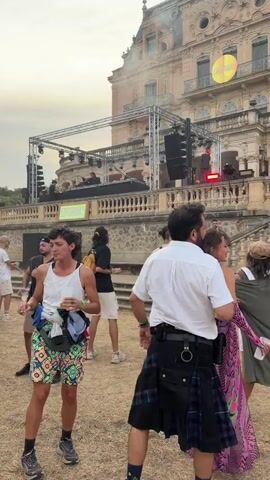 The video shows a group of people gathered outdoors in front of a large, ornate building that appears to be a historic mansion or chateau. There is a stage set up with lighting and speakers, suggesting a performance or event is taking place. Several people are visible in the foreground, some dancing and others observing the stage. One person in the foreground wears a white shirt and a kilt, another wears a white tank top and patterned shorts, and a third wears a vibrant pink and purple patterned dress. The building has multiple levels, balconies, and a domed roof. The grounds appear to be made of dirt and gravel. The sky is overcast, suggesting it might be late afternoon or early evening. The overall atmosphere seems festive and relaxed.