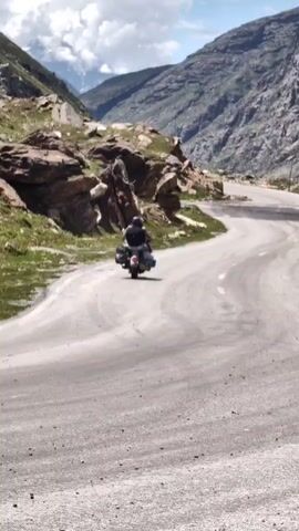 A motorcycle rider, seen from behind, travels along a winding paved road through a mountainous, rocky landscape. The rider is wearing a helmet and a backpack. The road curves to the right, with steep, rugged mountains on either side. Patches of green grass are visible on the slopes, and the sky is partly cloudy with some distant snow-capped peaks.