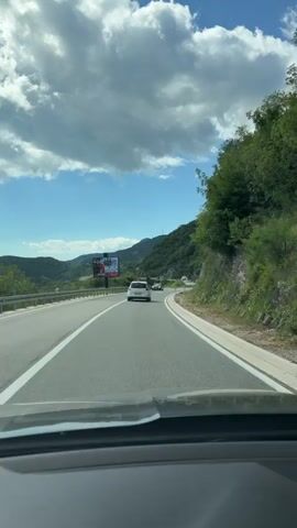 The video shows a view from inside a car driving on a two-lane road. The road is paved with white lines indicating the lanes. On the left side of the road, there is a green landscape with hills and trees. A billboard advertising a political campaign is visible in the distance. Several cars are on the road, some in front of and some passing the camera car. On the right side of the road, there is a steep, rocky hill covered with green vegetation. The sky is blue with scattered white clouds. The camera is angled slightly downward, showing the dashboard of the car. The footage appears to be taken during daylight.