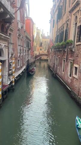 The video shows a canal in Venice, Italy. The water is green and reflects the sunlight. A gondola with a gondolier in black is navigating the canal. The buildings on either side of the canal are old and made of brick. There are flags hanging from some of the buildings. A small bridge arches over the canal in the distance. Another gondola is partially visible on the right side of the frame. There are people walking on the bridge. The overall scene is tranquil.