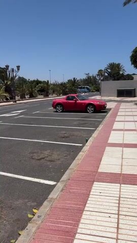A red convertible is parked in a parking lot. The car is facing the right side of the frame. In the background, there are palm trees and a blue bus. The sky is clear and blue.