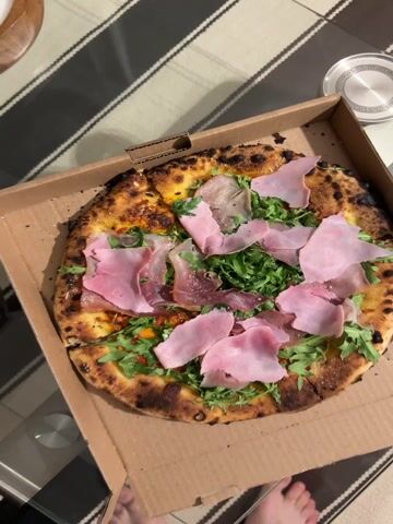 The video shows a pizza in a cardboard box. The pizza has a burnt-looking crust, a yellow base, green arugula, and slices of pink prosciutto on top. The camera zooms in on the pizza to show more detail. The background has a striped surface and a silver object in the corner.