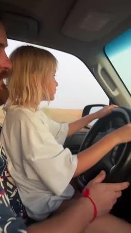 A young girl with blonde hair sits in the driver's seat of a car, holding the steering wheel. A man's arms are visible in the foreground, with one hand resting on the steering wheel near the girl's and another holding her left arm. The car appears to be moving, with the scenery outside blurred. The interior of the car includes a dashboard with a multimedia system. The exterior shows a dry, open landscape under a hazy sky, possibly during sunset.