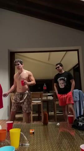 The video depicts a casual indoor setting where several individuals are gathered. Initially, two men are visible: one shirtless, wearing patterned shorts and sunglasses, holding a red cup, and the other in a black t-shirt, red shorts, and sunglasses. A third man, wearing a red robe, walks toward a white couch where two women are sitting. He offers a red cup to one of them. Behind them, a man in a patterned shirt stands with a drink. There are red, orange, yellow, and blue plastic cups on a glass-topped table along with a brown bottle and a cigarette pack. The room is illuminated, with a view of an outdoor area visible through a large glass door. The atmosphere seems relaxed.