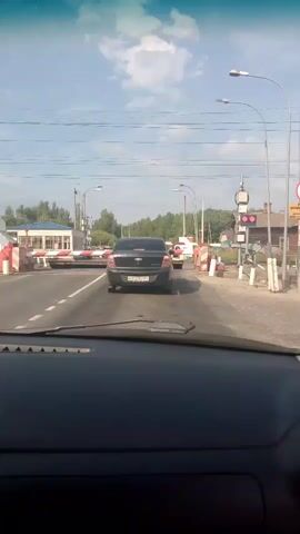 The video, captured from inside a vehicle, shows a view of a road leading to a railroad crossing. In the foreground, the dashboard and windshield of the car are visible. Ahead, a black sedan is stopped on the road, facing the crossing. The railroad crossing arms are down, indicating an approaching train or a train passing. There are other vehicles and structures around the crossing, including buildings and traffic lights. The sky is blue with some clouds. The overall scene depicts a typical road situation at a railroad crossing.