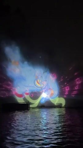 A nighttime video displays a projection onto water, possibly a mist or fog screen. The projection shows a large blue animated genie-like character with yellow accents. The character appears to be moving and interacting with the projected environment. In the foreground, the surface of the water reflects the projected light. There is a dark, flat structure in the lower left corner of the frame, possibly a dock or platform. The sky is dark and obscured. The video captures the projected animation in segments, showcasing different poses and movements of the blue character.