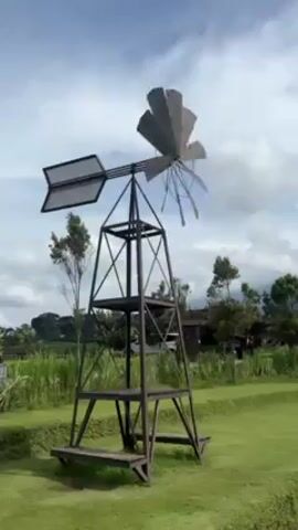 The video shows a decorative windmill rotating on a sunny day. The windmill is made of dark metal and stands on a tiered base. The blades of the windmill are spinning gently. The background consists of a green lawn, trees, and a partly cloudy sky. As the video pans, people can be seen walking along a path in the background, and a clock tower is visible. The landscape is slightly terraced, creating a patterned effect on the lawn.