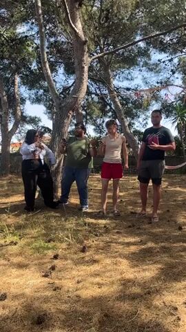 Four people stand under a tree in a yard. The yard is covered in dry grass and pine cones. From left to right, the first person is wearing a white shirt, black pants, and sunglasses. The second person is wearing an olive green shirt and blue jeans. The third person is wearing a white tank top and red shorts. The fourth person is wearing a black shirt and green shorts. All four people are holding cell phones and appear to be singing or talking. In the background, there are other trees, a fence, and a hammock. The sky is blue.