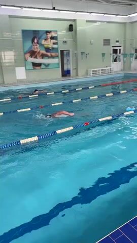 An indoor swimming pool with multiple lanes marked by blue and white buoys. Several individuals are swimming in different lanes. In the foreground, a person in an orange swimsuit is swimming freestyle, with water splashing around them. In the background, another person is swimming in a different lane, and further back, a third person can be seen. The pool area has tiled walls and a high ceiling with fluorescent lighting. A large poster with images of two women exercising is visible on the wall. The water is a clear blue color.