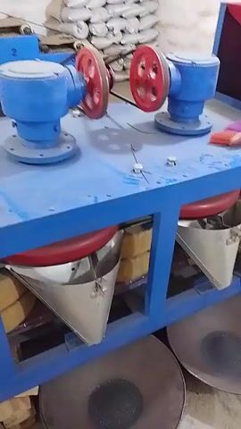 The video shows a machine with rotating blue and red components on a blue frame. Underneath are two metal funnels feeding into two circular pans containing a dark material. The machine appears to be processing something, possibly separating or mixing materials. The environment looks like a workshop or industrial setting, with a concrete floor.