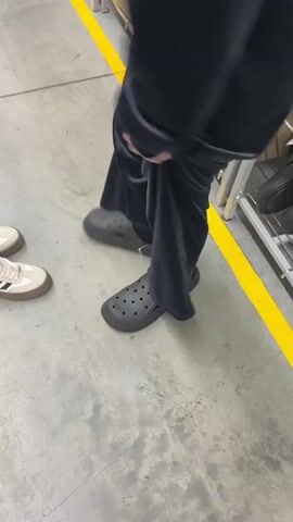 The video shows a person from the waist down, wearing black velvet-like pants and black Crocs with black socks. The person is standing on a concrete floor next to a yellow safety line. A pair of light-colored Adidas sneakers are on the floor to the left of the person. Behind the person, there's a metal shelving unit filled with cardboard boxes. The person lifts their pants slightly, revealing more of the Crocs. The camera is held at a low angle and remains mostly stationary throughout the short clip.