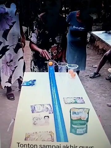 A ping pong ball rolls down a blue ramp on a white table. A person is leaning over the table, apparently blowing on the ball to move it. There are two clear plastic cups on the table, one of which contains ping pong balls. There are also various products and wrappers on the table, including "Giv" soap and a "Mama" product. Several people are standing around the table, observing the game. The environment appears to be outdoors, with dirt and concrete visible on the ground. The text at the bottom of the screen reads "Tonton sampai akhir guys..".