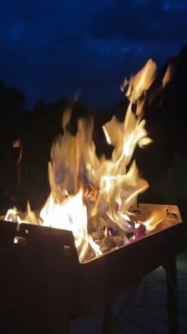 The video shows a close-up view of a campfire at dusk. Flames flicker and dance in a metal fire pit, casting a warm glow against the dark night sky. The fire pit is situated outdoors, with trees and hills visible in the background. The overall atmosphere is serene and tranquil.