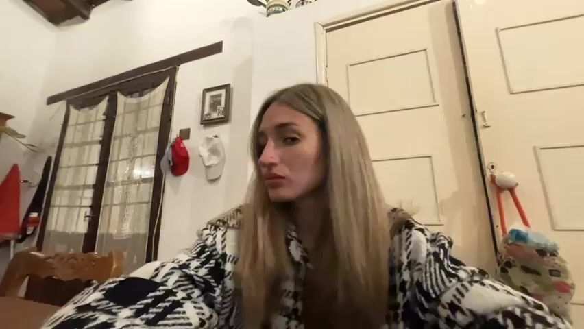 A woman with long blonde hair is looking at the camera and moving her head. She is wearing a black and white patterned jacket. The room is dimly lit and appears to be a living room. There are curtains in the background and a door on the right side of the frame.