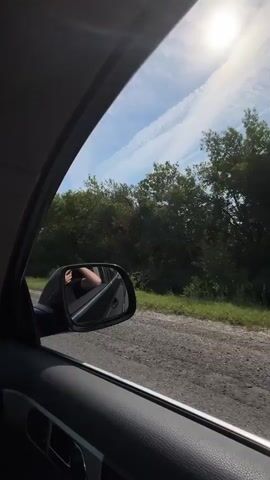 The video shows a view from inside a car looking out the passenger window. The car is driving on a dirt road next to a paved road. Trees and bushes line the side of the road. The sun is shining brightly in the sky, and there are some clouds. In the distance, on the paved road, there are two red trucks driving ahead. As the car continues, it merges onto the paved road behind the trucks. The camera then pans forward, showing a large white trailer truck passing the camera on the left. The two red trucks are still visible ahead of the camera. The video appears to be shot on a sunny day.