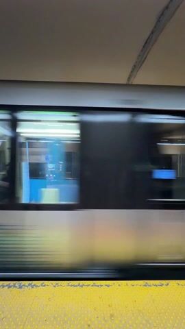 A subway train moving past the camera on a platform in an underground station. The train is mostly blue with a white horizontal stripe. The windows are dark and tinted, but silhouettes of people can be seen sitting and standing inside. The platform has a yellow safety line with raised dots. The station ceiling is a light beige. The train is moving fast, creating a blurring effect.
