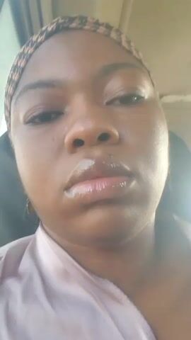 The video shows a close-up of a young Black woman from the shoulders up. She is wearing a patterned headband and a light-colored robe or shirt. Her eyes are closed for the first few seconds, then they open. The lighting is bright and appears to be from an outdoor source, possibly sunlight. The background is blurred and out of focus, but suggests an indoor environment, possibly a car. There is a bright light source to the right of the frame, causing some glare. The woman appears to be looking at the camera.