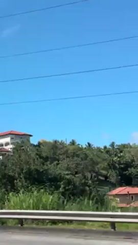 From a low angle, a car drives on a road. On the right, there is a metal barrier. Behind the barrier, there are many green leafy trees. On top of a hill behind the trees, there is a multi-story house with a red-tiled roof. The sky is bright blue with no clouds. Power lines run across the top of the scene. The car passes another white SUV on the road.