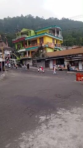 A short video shows a street scene in a village with a colorful building in the background. Children are playing on the street. Some are riding small vehicles, possibly scooters or tricycles. A man walks across the street. A motorbike passes by. The building in the background is multi-story and painted in bright colors like yellow and green. There are trees and vegetation on the hillside behind the building. The sky appears overcast. The street is paved with asphalt. There are some parked vehicles and small shops or stalls along the street. The overall impression is a lively village street with children engaged in play.