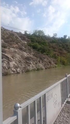 A wide shot shows a river flowing past a rocky hill. In the foreground, a metal railing runs along the edge of a paved area next to the river. The railing has a sign on it that reads "BULOOQ HILLS." The hill is covered in rocks and sparse vegetation. The river is wide and murky, with some ripples on the surface. The sky above is blue with some scattered clouds. The camera pans slowly to the right.