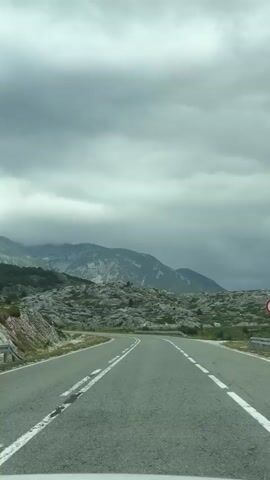 A car travels on a winding road through a mountainous landscape. The road is paved and has white lane markings. The surrounding terrain is rocky with sparse vegetation. Mountains are visible in the background, partially obscured by clouds. The sky is overcast.