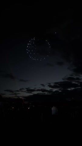 The video shows a nighttime scene with what appears to be a drone light show in the sky. Many lights are arranged in a circular shape, resembling a moon. The sky is dark, with some clouds visible in the background near the horizon. In the foreground, there's a silhouette of people watching the show.