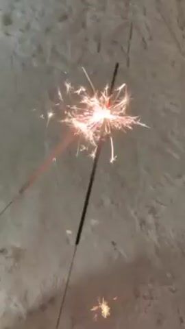 A hand with red nail polish holds a lit sparkler, which is emitting bright sparks. The sparkler is against a light gray, textured surface. The camera angle shifts to show a patch of snow, where a separate, lit sparkler lies on the ground. Another hand, wearing a yellow sleeve, moves in the frame near the sparklers. The primary focus remains on the sparkler being held, with the ground sparkler occasionally appearing in the background.