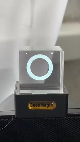 The video displays a digital animation of a face projected onto a clear cube, which rests on a black base with a yellow "GeekMagic" label. The projected face features simple white eyes and a mouth that changes expressions. The animation cycles through a few different mouth shapes, including a wavy line, a wide smile, and a circular shape. The background is blurred but appears to be indoors with soft, diffused lighting, possibly from a window. The overall mood is lighthearted and playful due to the animated face. The text "GeekMagic" is clearly visible on the base of the device. The setting is indoors, likely a home or office. There are no people visible in the video. The time of day and weather are not discernible.