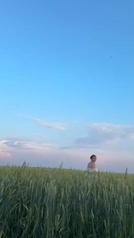 A person wearing a white off-the-shoulder top is sitting in a field of tall green grass. The person turns their head to the left, then to the right. They then raise their arms to the sky. The sky is blue with scattered white clouds and a pinkish horizon.