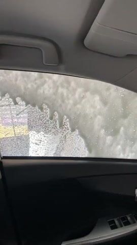 The video shows the interior of a car. The view is through the passenger window, looking outwards, with some suds or foam obscuring the view. The dashboard and part of the steering wheel are visible. A rosary hangs from the rearview mirror. The car appears to be in motion, possibly at a car wash.