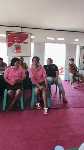 The video shows a group of people sitting in a room with a red carpet and white walls. There are windows and doors in the background. Some people are sitting in chairs, while others are standing. There are tables and chairs set up in front of some of the people. There are also posters on the walls. Some of the people appear to be listening to someone speak, while others are engaged in conversation. There is a water bottle and a microphone in the foreground.