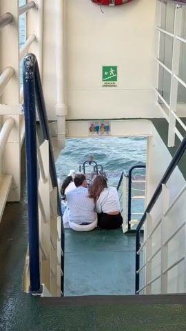 The video shows a group of people sitting on the deck of a ferry near an open doorway leading to the water. A man and a woman are sitting closely together, facing away from the camera towards the water. The man is wearing a striped shirt and white pants, and the woman is wearing a white top and dark pants. Another person, possibly a child, is visible between them, wearing white. They are seated on a green surface, and a metal railing is visible to the left and right of the frame. In the background, there is a white wall with a green emergency exit sign depicting a person running down stairs. The water is visible through the open doorway, appearing choppy. There is a red life preserver visible on the wall above the doorway. The overall environment appears to be the exterior deck of a ferry.