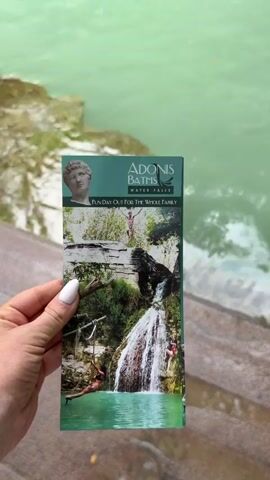 The video shows a person holding a brochure for Adonis Baths Water Falls, and panning up to show the location advertised on the brochure. The Adonis Baths Water Falls is a natural pool with a small waterfall emptying into it. There are some people swimming in the pool. The surrounding area is lush and green, with trees and plants growing on the rocks around the pool. The water is a clear turquoise color.