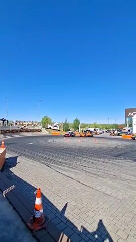 The video captures a drift racing event outdoors under a clear blue sky. Several cars are actively drifting in a circular track marked by orange cones. One car, painted yellow with racing decals, is particularly visible performing a drift, emitting smoke from its tires. Spectators are lined up along the perimeter of the track, behind barriers. The track itself is paved with interlocking bricks. In the background, there are buildings and trees, suggesting an urban or suburban setting. The overall atmosphere appears to be one of excitement and skilled driving.