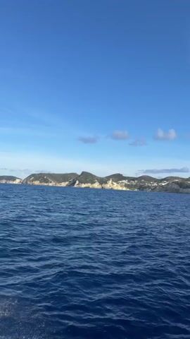 A boat is moving through the ocean, creating a wake. The water is a deep blue, and the sky is a clear blue with a few white clouds. In the distance, there is a green island with white buildings on it. The island has a rugged coastline. The video is shot from the perspective of someone on the boat.
