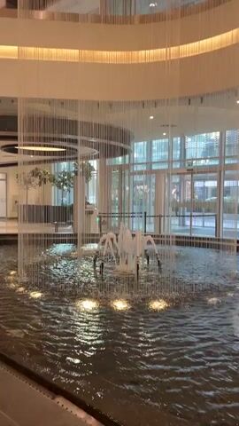 The media file shows an indoor fountain with a water curtain feature in a modern shopping mall. The fountain is centrally located in a large open space with multiple levels of balconies visible above. The water curtain is made of numerous thin strands that water cascades down, creating a shimmering effect. The fountain itself has several jets spraying water upwards. The lighting is bright and artificial, suggesting an indoor environment during daylight hours or a well-lit evening. The architecture of the mall is contemporary, with clean lines and polished surfaces. The overall atmosphere appears calm and serene, highlighting the visual appeal of the water feature. There are no people clearly visible in the frame, but the presence of a security camera at the top left suggests a public space. The location context provided, Ataşehir, Turkey, indicates the geographical setting of this scene. The mall's interior design, with its circular elements and light-colored finishes, contributes to a sense of spaciousness and elegance. The presence of trees on one of the balconies adds a touch of nature to the man-made environment.