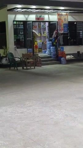 The video shows the exterior of a small store at night. The store, called “BUNIMART”, has an open door, revealing shelves stocked with goods. There are large bottles of water stacked on the steps leading up to the entrance. In front of the store, there are a few chairs. To the right, a black car is parked. The area is well-lit, but the sky is dark. There is some indistinct audio of people speaking. The camera pans slightly from left to right and back again, showing the entire scene. There is a “BABLISS” sign hanging on the store.