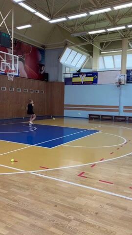 A female is practicing tennis in an indoor gymnasium. The woman is fair-skinned and blonde, wearing a black athletic dress and white tennis shoes. She is holding a tennis racket and hitting yellow tennis balls that are scattered on the floor. She moves around the blue and yellow basketball court, hitting the balls with forehand strokes.  The gym has a polished wooden floor and a high ceiling with skylights. A large basketball hoop is visible on the left side of the frame.  There are wooden benches along the wall and an image of a basketball player on the wall.  Banners with text in Russian hang on the wall above the benches.