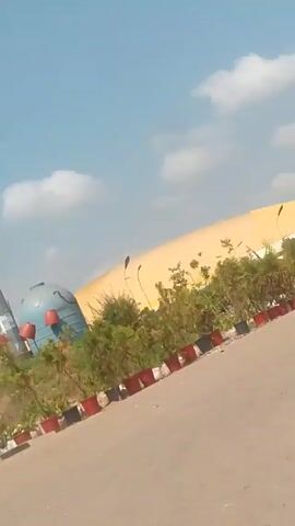 The video, taken from a moving vehicle, shows a roadside scene on a bright, sunny day. There are various structures including a tall, cylindrical pole, a low wall with flowerpots, and a yellow building with an awning. There is also a large, metal electrical transmission tower. A blue and white Nissan van, with a red cross, is parked near the roadside, and people are standing beside it under a blue awning. The video is somewhat shaky and the view is partially obstructed.
